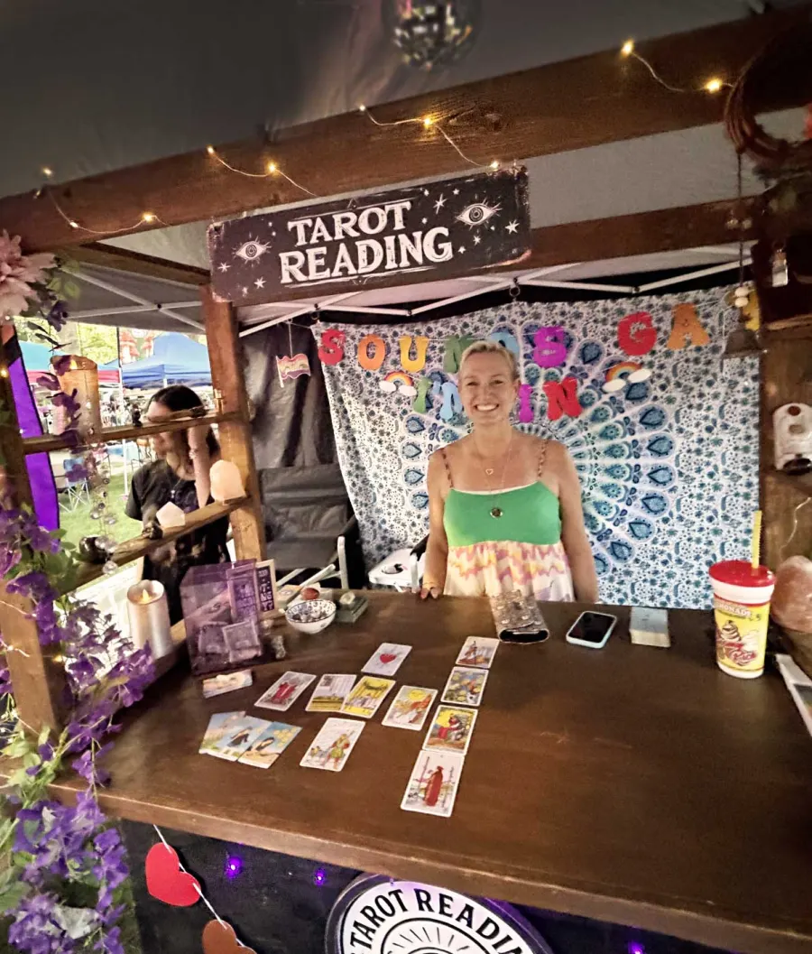 Mamma T. smiling behind her wooden booth, with tarot cards laid out on the table