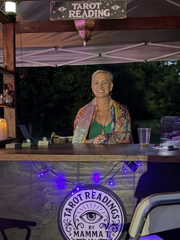 Mamma T. smiling behind her booth in the evening, under a Tarot Reading sign